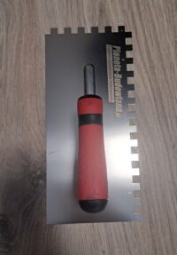 Paca nierdzewna do gipsu PREFIX 127128E10-2K gryf aluminiowy, uchwyt dwukomponentowy, ząb 10/10 mm 280 mm (127128E10-2KO) Notched tile trowel with a red-and-black handle resting on a metal ruler used for spreading tile adhesive.