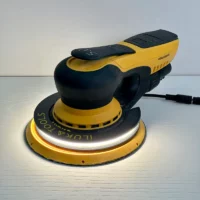 Corded yellow and black orbital sander resting on a white work surface, showing a circular sanding pad and glow from the pad/LED ring.